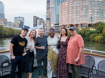 Six people smiling on a boat deck with urban skyline and river in background during daytime.