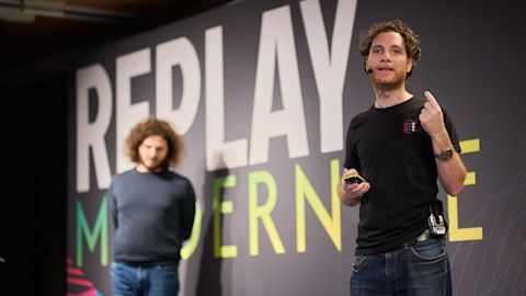 Man in black shirt presenting on stage with microphone headset, large text display behind him, another person visible.