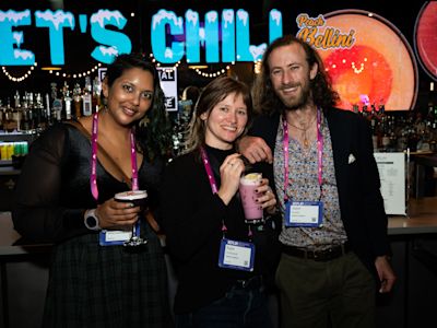 Three smiling people wearing name badges hold colorful drinks at a bar with neon signs reading Let's Chill in background.