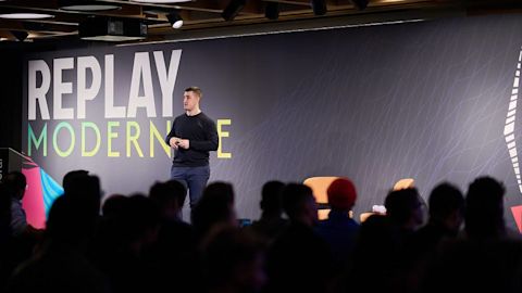 Speaker presenting on stage before an audience with large gray backdrop displaying "REPLAY MODERNIZE" text in white and green.