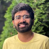 Smiling man with glasses and beard wearing yellow shirt against blurred green foliage background