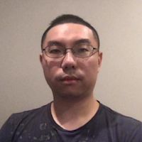 Asian man with short dark hair and glasses wearing a dark patterned t-shirt against a beige wall