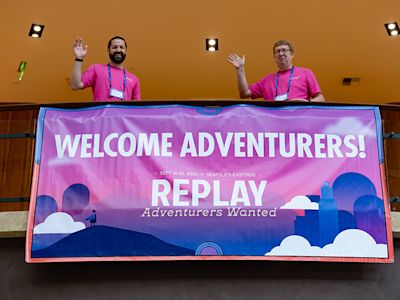 Two people in pink shirts waving behind a purple banner reading "Welcome Adventurers! REPLAY Adventurers Wanted"