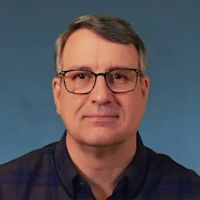 Professional headshot of a man with dark hair wearing black-framed glasses and a dark blue polo shirt against a blue background.