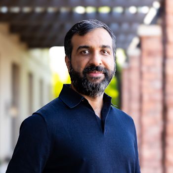 Samar Abbas, Man with dark hair and beard wearing navy blue polo shirt smiling at camera with blurred architectural background