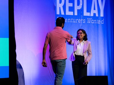 Two people on stage with blue backdrop displaying REPLAY Adventurers Wanted text, illuminated by purple and blue lighting.