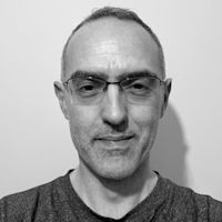 Black and white portrait of a man with short hair, wearing glasses and a dark t-shirt, looking directly at the camera.