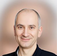 Professional headshot of a smiling man with short gray hair wearing a dark collared shirt against a soft beige background.