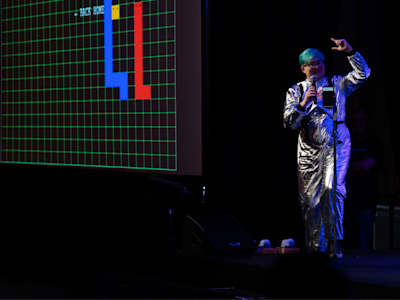 Performer in silver jumpsuit holding microphone on stage with retro video game screen displaying colorful blocks behind them