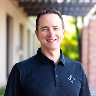 Man in navy blue polo shirt with white logo smiling at camera outdoors with blurred building background.