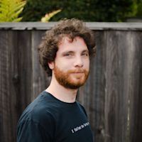 Man with curly brown hair and red beard wearing black t-shirt, smiling outdoors against wooden fence with greenery.