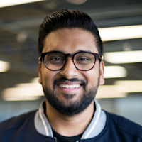 Smiling man with dark hair, beard, and black-framed glasses wearing a navy polo shirt in an office setting.