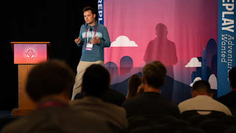 Speaker presenting to an audience on stage with pink backdrop featuring clouds and city skyline graphics.