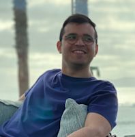 Smiling man wearing glasses and a navy blue shirt, holding a green striped pillow, seated indoors with blurred background.