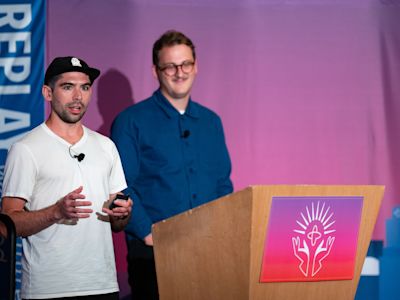 Two men presenting at a podium with a colorful logo, one speaking with microphone, against a purple and pink background.