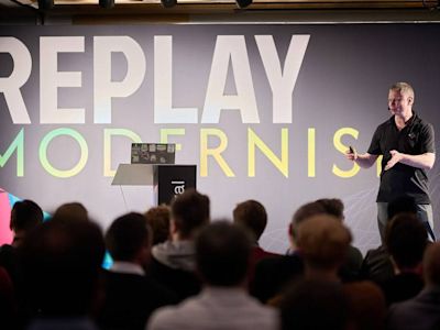 Speaker in black shirt presenting to audience with large REPLAY MODERNIST sign on gray backdrop behind podium.