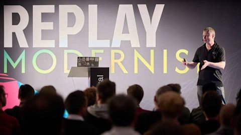 Speaker in black shirt presenting to audience with large REPLAY MODERNIST sign on gray backdrop behind podium.