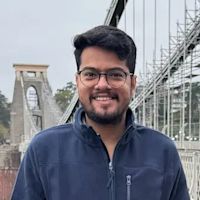 Young man with glasses and dark hair wearing a navy blue fleece jacket, smiling at camera with a suspension bridge in background.