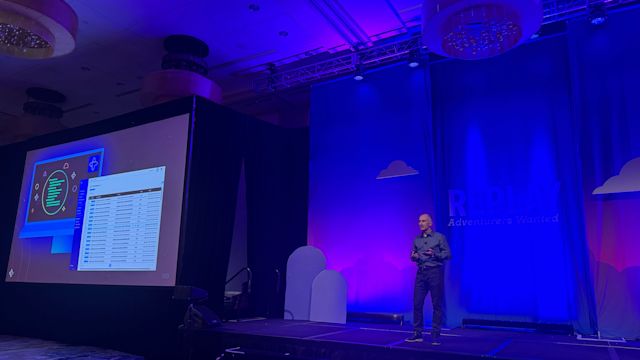 Speaker presenting on stage in blue-lit ballroom with large screen displaying technical diagrams and data spreadsheet.