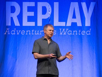 Man in gray shirt presenting on stage with blue backdrop displaying REPLAY Adventures Wanted text