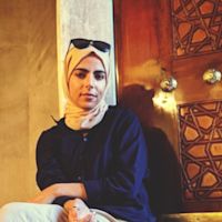 Woman wearing yellow hijab and sunglasses on head, dressed in black, seated indoors with decorative wooden screen behind her.