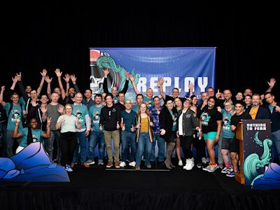 Large group of people in teal shirts posing on stage with raised hands under a blue banner with octopus graphics.