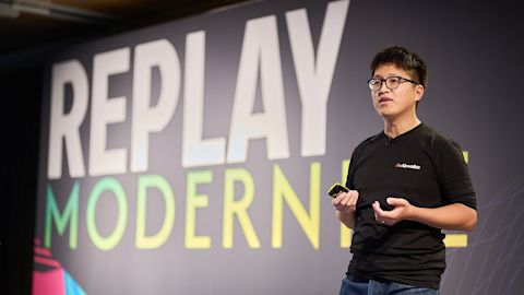 Person in black shirt and glasses presenting in front of large screen displaying REPLAY MODERN in white and green text.