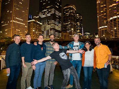 Group of eight people posing together at night with illuminated Chicago skyscrapers in the background along the river.
