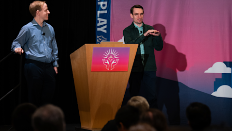 Two men presenting on stage at a podium with a colorful logo, one gesturing while speaking to an audience below.
