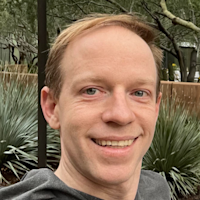 Smiling man with light brown hair in gray shirt outdoors with bare trees and green plants in background.