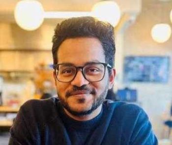 Naveen Achyuta, Smiling man with glasses and beard wearing dark clothing in a bright indoor space with round lights in background