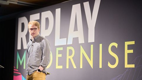 Young man with glasses wearing checkered shirt presenting on stage with large screen displaying text behind him.