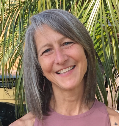 Smiling woman with shoulder-length gray hair wearing a mauve shirt, photographed outdoors with green palm fronds in background.