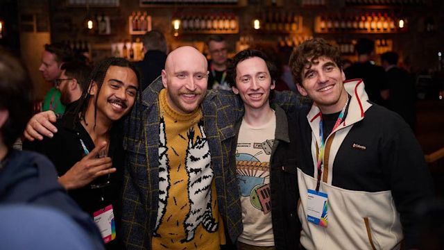 Four smiling friends pose together at a bar with bottles displayed on shelves in the background.