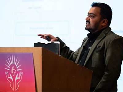 Man in olive green jacket speaking at wooden podium with purple and pink logo, gesturing with remote in hand.