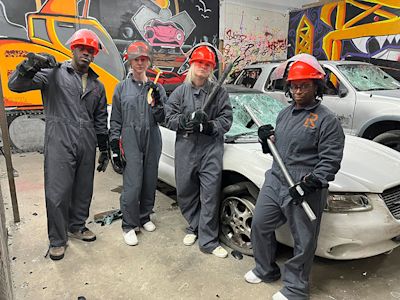 Four students in gray coveralls and red hard hats stand with tools near damaged cars and colorful graffiti murals.