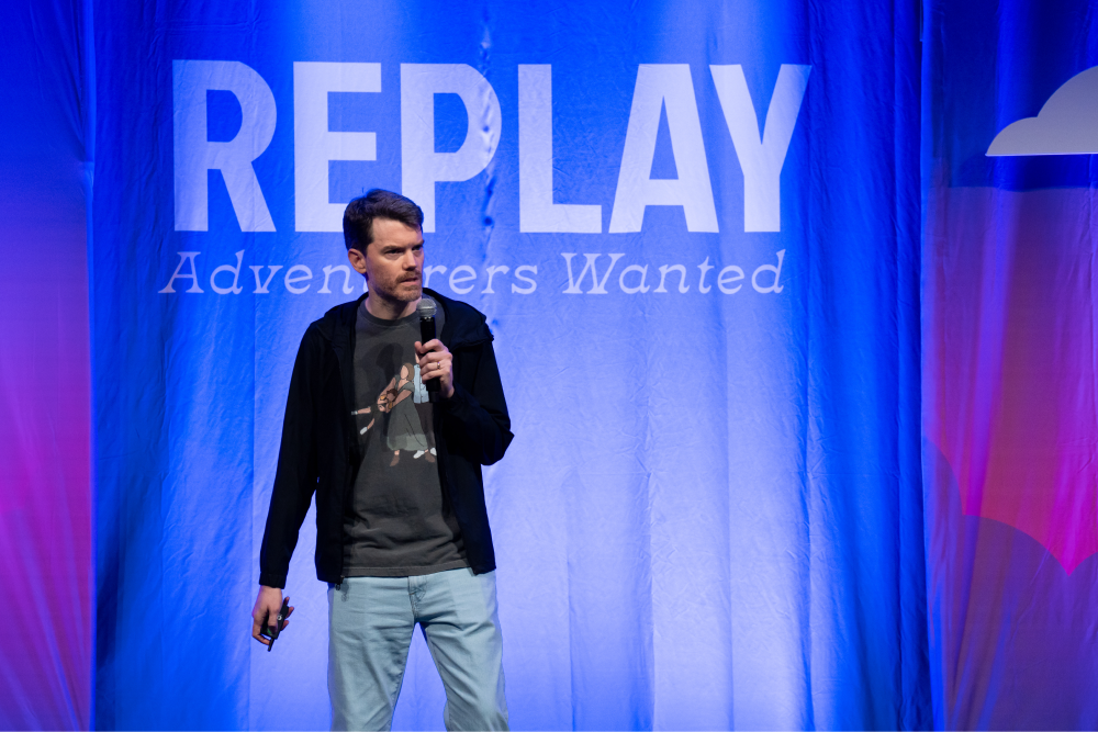 Speaker with microphone on stage in front of blue backdrop displaying REPLAY Adventurers Wanted text