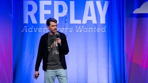 Speaker with microphone on stage in front of blue backdrop displaying REPLAY Adventurers Wanted text