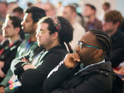 Diverse audience members listening attentively at an event, with a man in glasses and cornrows in focus in the foreground.