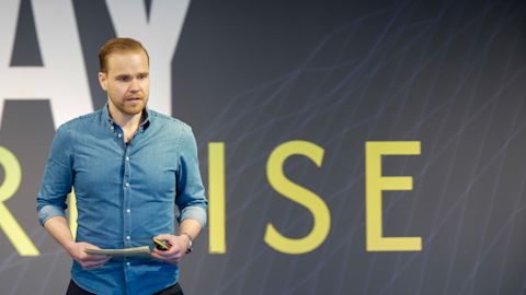Man in blue denim shirt holding papers and speaking at presentation with yellow text on dark background