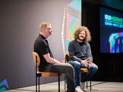 Two men seated on wooden chairs on stage having a conversation, with colorful projected graphics on screens behind them.