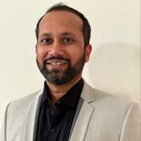 Professional headshot of a smiling man with a beard wearing a light gray blazer and black shirt against a white wall.