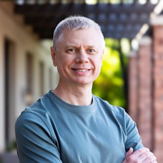 Middle-aged man with short gray hair smiling at camera, wearing a blue-gray t-shirt with arms crossed, outdoors.