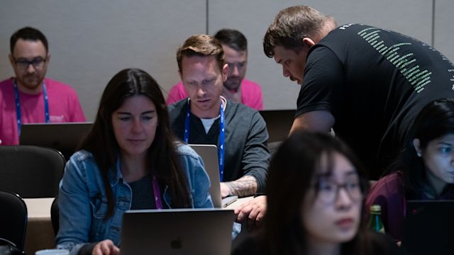 Group of people working on laptops at a tech event, with staff in pink shirts assisting participants with blue lanyards.