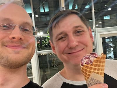 Two men taking a selfie indoors, one wearing glasses, the other holding an ice cream cone with purple topping.