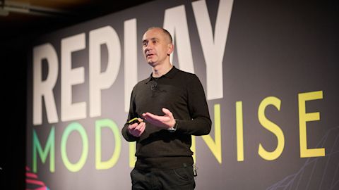 Man in black sweater presenting on stage with large screen displaying text REPLAY and MODERNISE behind him