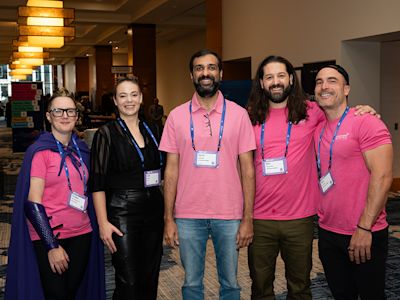 Five conference attendees wearing pink shirts and blue lanyards pose together in a hotel lobby with warm lighting.