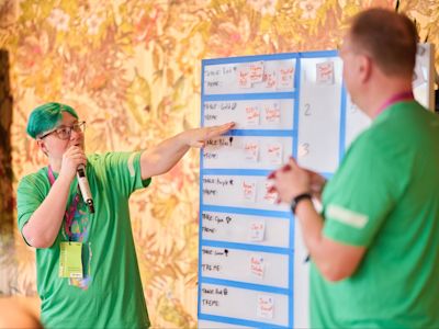 Person with green hair and glasses in green shirt pointing at whiteboard with table assignments during presentation