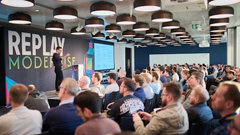 Speaker presenting to large audience in modern conference room with round pendant lights and Replay Modernise branding.