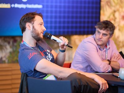 Man with beard speaking into microphone at panel discussion while another man in pink shirt listens at table.